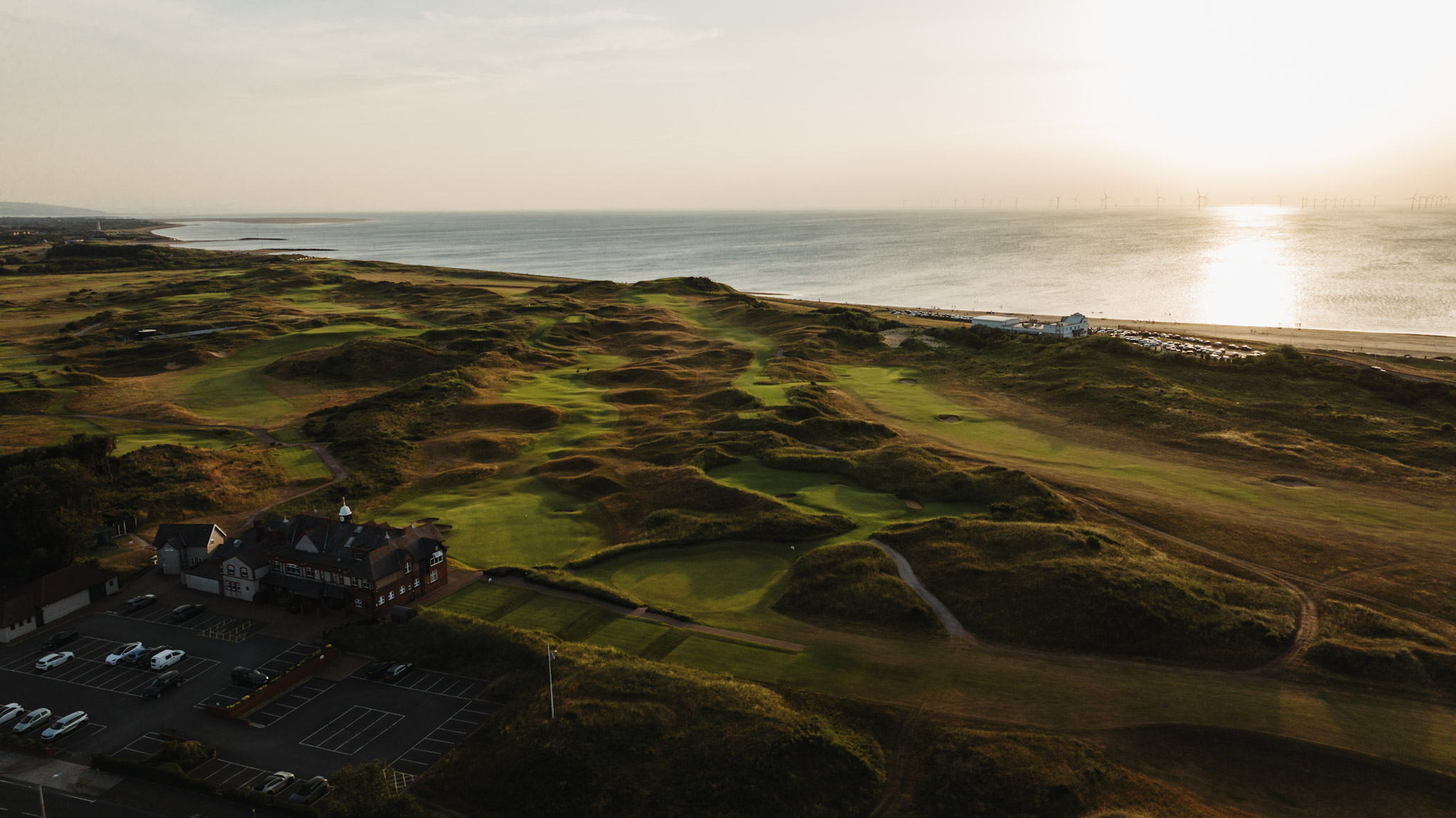 The clubhouse and car park from the air at sunset, the links running out to the sea and the wind farm on the horizon