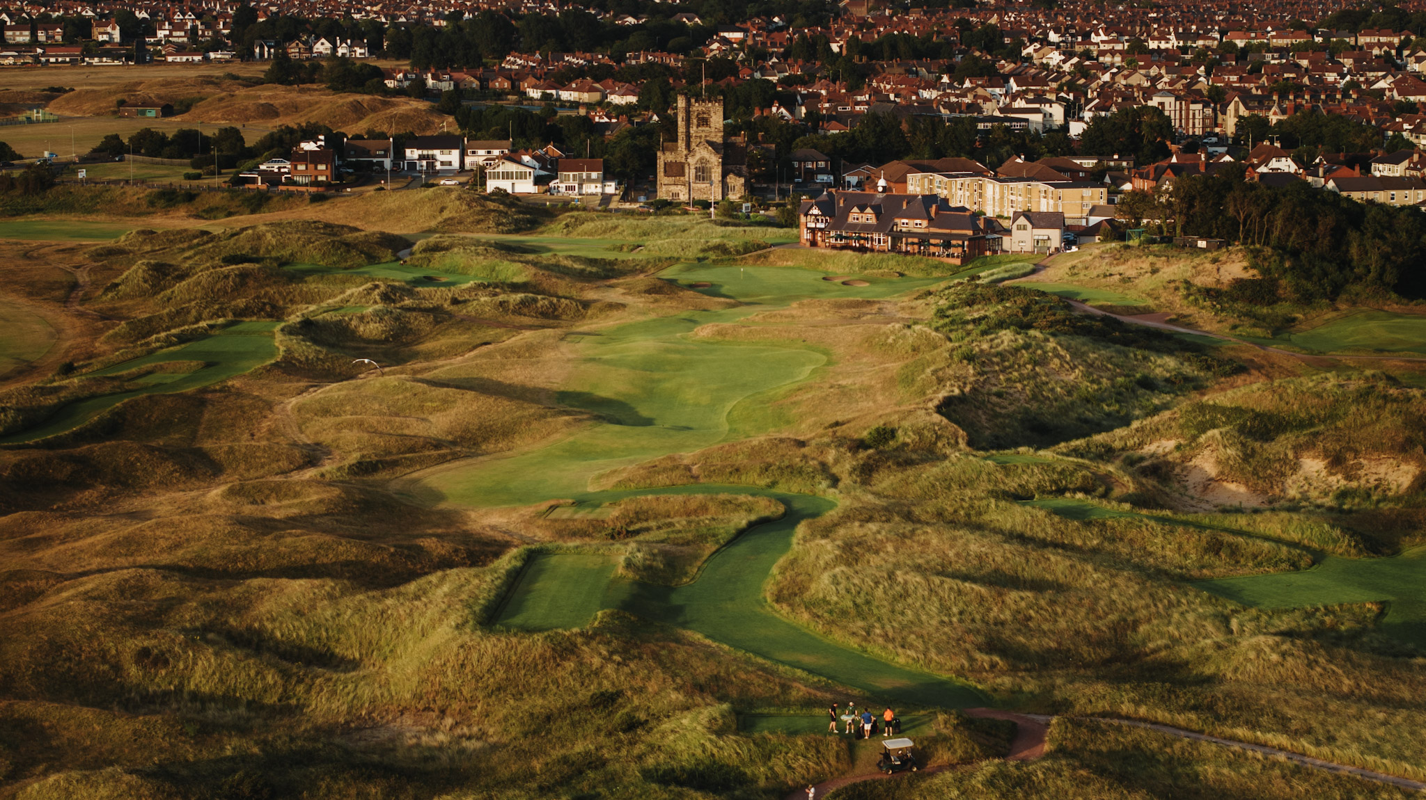 A fourball on the tee from the air, the fairway winding through the dunes up to the clubhouse and the church tower beyond